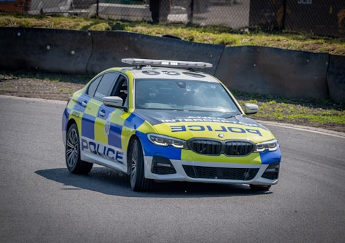 A police car with a blue and yellow checkered pattern is parked on a paved road near a grassy embankment. The vehicle has the word 'POLICE' prominently displayed on the side, and it features a roof-mounted light bar. The background includes a barrier and some industrial equipment.