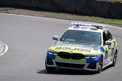 A marked police interceptor vehicle with visible blue and yellow checkered patterns is driving on a curved, paved road. The car has a light bar on top and police markings in bold letters on the hood and sides.