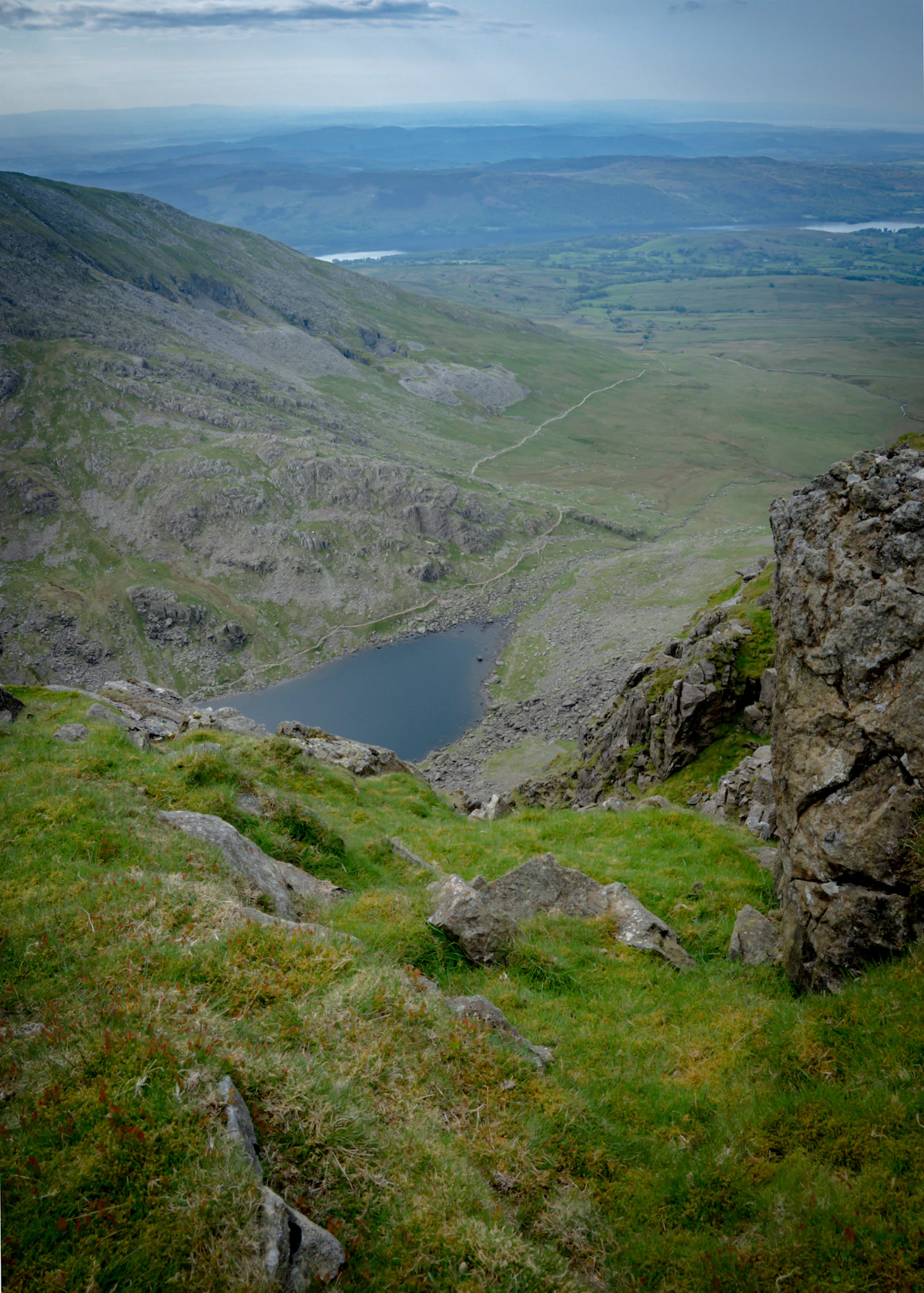 A tarn below rocky slopes on the Old Man of Coniston, with lakes and low hills beyond