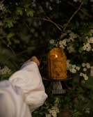 A hand in a white sleeve holds an ornate, partially melted yellow candle on a rustic candleholder. The background consists of green foliage with white flowers, suggesting a natural, outdoor setting.