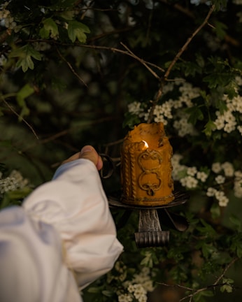 A hand in a white sleeve holds an ornate, partially melted yellow candle on a rustic candleholder. The background consists of green foliage with white flowers, suggesting a natural, outdoor setting.