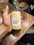 A hand is holding a glass bottle of sparkling kombucha labeled 'umami brew' with a 'Pineapple Spice' flavor. The bottle is displayed against a wooden table surface. A glass filled with a beverage is visible in the background.