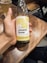A hand is holding a glass bottle of sparkling kombucha labeled 'umami brew' with a 'Pineapple Spice' flavor. The bottle is displayed against a wooden table surface. A glass filled with a beverage is visible in the background.