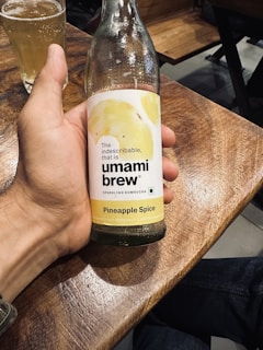 A hand is holding a glass bottle of sparkling kombucha labeled 'umami brew' with a 'Pineapple Spice' flavor. The bottle is displayed against a wooden table surface. A glass filled with a beverage is visible in the background.