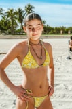 A person stands confidently on a sandy beach, wearing a yellow floral bikini and a black bead necklace. In the background, tall palm trees are visible under a clear blue sky. The warm sunlight casts shadows on the sand, creating a bright and inviting atmosphere.