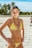 A person stands confidently on a sandy beach, wearing a yellow floral bikini and a black bead necklace. In the background, tall palm trees are visible under a clear blue sky. The warm sunlight casts shadows on the sand, creating a bright and inviting atmosphere.