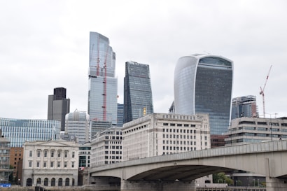 A cityscape featuring modern skyscrapers with unique architectural designs, juxtaposed with classical buildings in the foreground. A bridge extends across the bottom part of the image, adding depth to the urban scene. The sky is overcast, contributing to a muted yet sophisticated atmosphere.