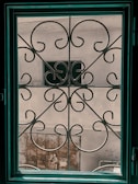 A decorative iron grill with ornate swirling patterns is seen through a window. The window frame is dark teal and contrasts with the neutral tones of the wall outside. There is a weathered metal gate and a partial view of another structure in the background.