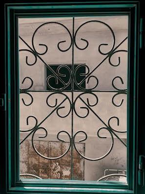 A decorative iron grill with ornate swirling patterns is seen through a window. The window frame is dark teal and contrasts with the neutral tones of the wall outside. There is a weathered metal gate and a partial view of another structure in the background.