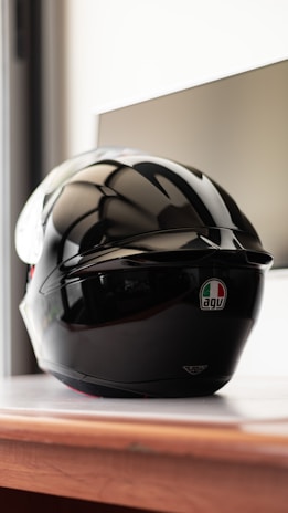 A glossy black motorcycle helmet with an AGV logo sits on a wooden surface. The background is softly blurred, highlighting the helmet's sleek and polished design.