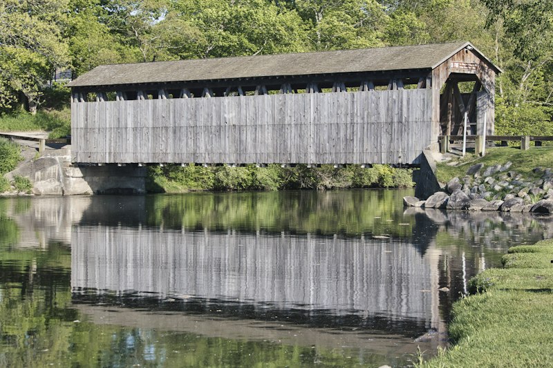A weathered gray wooden covered bridge reflected in a calm river on a sunny day