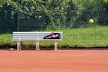 A tennis court with red clay surface, featuring a white bench with a sports bag placed on it. A tennis ball is captured in mid-air, and a chain-link fence with green grass and foliage is in the background.