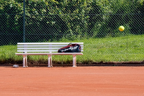 A tennis court with red clay surface, featuring a white bench with a sports bag placed on it. A tennis ball is captured in mid-air, and a chain-link fence with green grass and foliage is in the background.