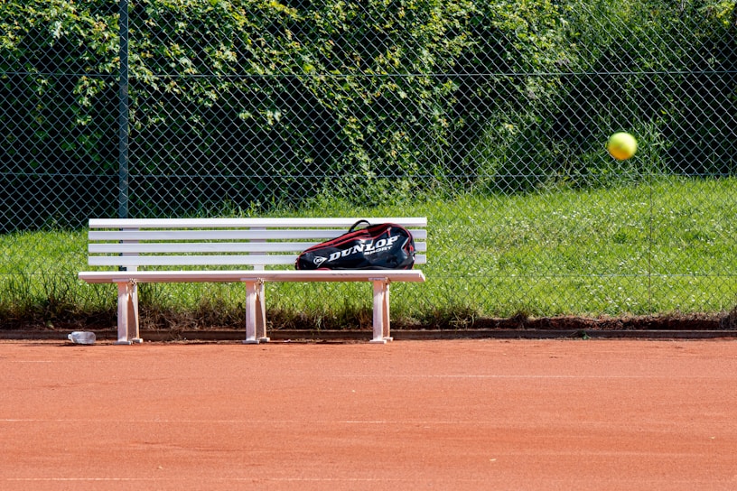 A tennis court with red clay surface, featuring a white bench with a sports bag placed on it. A tennis ball is captured in mid-air, and a chain-link fence with green grass and foliage is in the background.