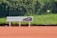 A tennis court with red clay surface, featuring a white bench with a sports bag placed on it. A tennis ball is captured in mid-air, and a chain-link fence with green grass and foliage is in the background.
