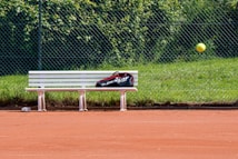 A tennis court with red clay surface, featuring a white bench with a sports bag placed on it. A tennis ball is captured in mid-air, and a chain-link fence with green grass and foliage is in the background.