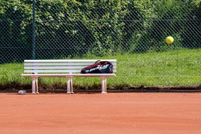 A tennis court with red clay surface, featuring a white bench with a sports bag placed on it. A tennis ball is captured in mid-air, and a chain-link fence with green grass and foliage is in the background.