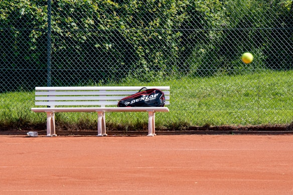 A tennis court with red clay surface, featuring a white bench with a sports bag placed on it. A tennis ball is captured in mid-air, and a chain-link fence with green grass and foliage is in the background.