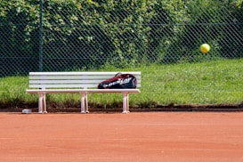 A tennis court with red clay surface, featuring a white bench with a sports bag placed on it. A tennis ball is captured in mid-air, and a chain-link fence with green grass and foliage is in the background.