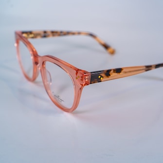 A pair of eyeglasses with transparent pink frames and tortoiseshell-patterned arms is placed on a light surface. The lenses are clear, and the arms are partially translucent with a mix of brown and black spots.