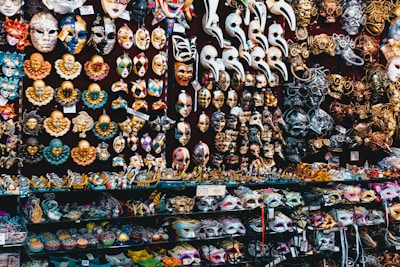 A vibrant display of Venetian masks fills the entire scene, featuring various designs, colors, and expressions. The masks include traditional faces, clowns, and elaborate creations adorned with gold, silver, and multicolored patterns. Some have intricate detailing with beads and feathers. The background is densely packed with shelves showcasing an assortment of masks and small figurines.