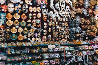 A vibrant display of Venetian masks fills the entire scene, featuring various designs, colors, and expressions. The masks include traditional faces, clowns, and elaborate creations adorned with gold, silver, and multicolored patterns. Some have intricate detailing with beads and feathers. The background is densely packed with shelves showcasing an assortment of masks and small figurines.