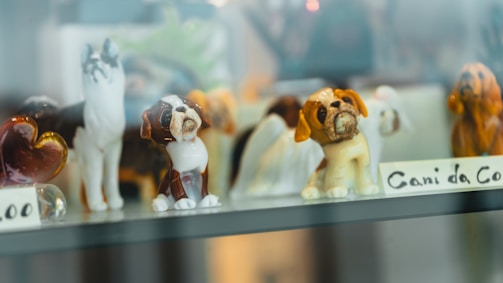 A collection of small ceramic or glass figurines displayed in a glass cabinet, including various small dog figures. The figurines are detailed, showing different breeds or styles and are set against a soft, blurred background.