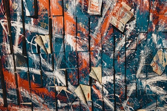 An abstract artwork composed of overlapping squares displaying a mix of bold, colorful streaks and splatters. The dominant colors are deep blue, bright orange, and white, with occasional pieces of paper, possibly from newspapers or books, scattered throughout the composition. These elements create a dynamic and chaotic visual effect.