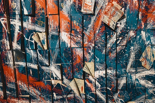 An abstract artwork composed of overlapping squares displaying a mix of bold, colorful streaks and splatters. The dominant colors are deep blue, bright orange, and white, with occasional pieces of paper, possibly from newspapers or books, scattered throughout the composition. These elements create a dynamic and chaotic visual effect.