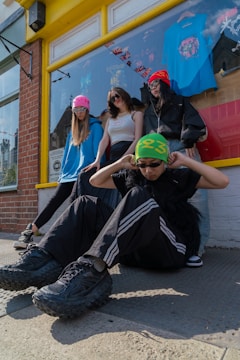 Four people are posed in front of a shop with a yellow frame around the window. They wear casual streetwear with vibrant beanies and sunglasses, conveying a modern urban style. The shop window displays a blue t-shirt and various decorations inside.