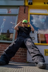 A person wearing oversized black cargo pants, a black shirt with textured embellishments, a neon green and yellow headscarf, and dark sunglasses stands with a confident pose in front of a brick building. Reflections in the glass show flags and clouds. The window displays a poster and the surroundings suggest an urban environment.