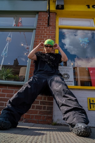 A person wearing oversized black cargo pants, a black shirt with textured embellishments, a neon green and yellow headscarf, and dark sunglasses stands with a confident pose in front of a brick building. Reflections in the glass show flags and clouds. The window displays a poster and the surroundings suggest an urban environment.