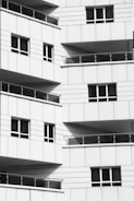 Multiple balconies with glass railings and a modern design style, featuring clean lines and grid patterns. The surface material appears to be a white or light-colored panel.