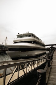 A large, multi-deck yacht is docked by a wooden pier, with the name 'Sir Winston' visible on its side. The background includes a glimpse of another vessel's masts and a bridge structure. The weather appears overcast, with the sky dominated by a pale gray tone.