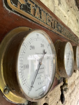 A set of vintage pressure gauges mounted on a wooden board with the label 'TANGYE' engraved above. The gauges feature intricate markings and measurements in feet head of water, indicating their use in measuring pressure. The brass casing around the gauges shows signs of aging and wear, adding to the antique appearance.