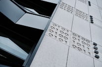 A close-up view of a modern building's facade featuring a grid of panels with embossed binary numbers (1s and 0s) and dark, rectangular windows. The angle of the photograph is tilted, creating a dynamic perspective.
