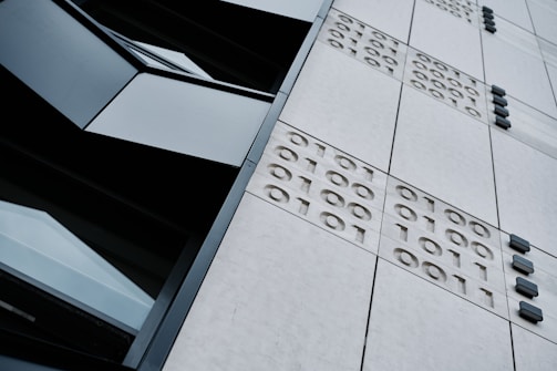 A close-up view of a modern building's facade featuring a grid of panels with embossed binary numbers (1s and 0s) and dark, rectangular windows. The angle of the photograph is tilted, creating a dynamic perspective.