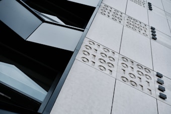 A close-up view of a modern building's facade featuring a grid of panels with embossed binary numbers (1s and 0s) and dark, rectangular windows. The angle of the photograph is tilted, creating a dynamic perspective.