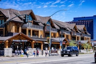 A rustic-style building featuring wooden architecture with several gables is situated along a city street. The building houses various businesses, including 'Powder House' and 'Zalanta'. People walk along the sidewalk, and a black SUV is parked on the street. A clear blue sky is visible in the background, along with a modern high-rise.