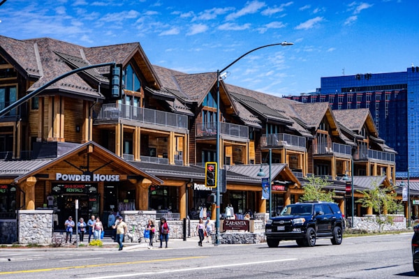 A rustic-style building featuring wooden architecture with several gables is situated along a city street. The building houses various businesses, including 'Powder House' and 'Zalanta'. People walk along the sidewalk, and a black SUV is parked on the street. A clear blue sky is visible in the background, along with a modern high-rise.