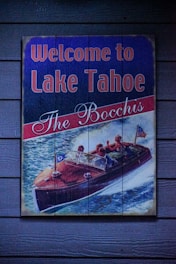 A vintage-style sign depicts a wooden speedboat on a body of water with people waving. The text reads 'Welcome to Lake Tahoe' and 'The Bocchis' in bold letters, set against a painted backdrop of blue water and white waves.