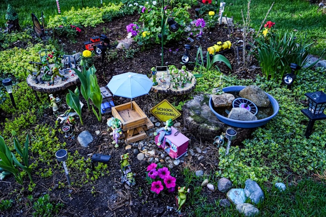 A whimsical garden setup featuring various decorative items, including small figurines of frogs, a tiny picnic table under a blue umbrella, a school crossing sign, and pastel-colored flowers. There are sections of green foliage, colorful flowers, and rocks arranged neatly within the garden.
