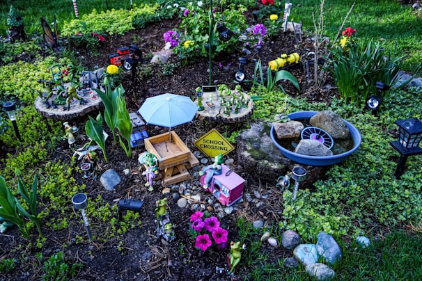 A whimsical garden setup featuring various decorative items, including small figurines of frogs, a tiny picnic table under a blue umbrella, a school crossing sign, and pastel-colored flowers. There are sections of green foliage, colorful flowers, and rocks arranged neatly within the garden.