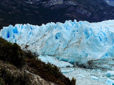 A vast glacier with jagged and towering ice formations dominates the landscape, surrounded by rugged mountainous terrain and some sparse greenery. The glacier appears to have varying shades of blue and white, indicative of compressed ice. The foreground shows a rocky outcrop with some vegetation, contrasting the cold ice with darker earthy hues.
