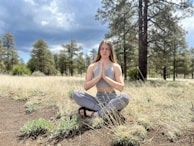 A person sits cross-legged on the ground with hands in a prayer position, surrounded by tall grass and pine trees. The sky is partly cloudy, creating a serene atmosphere.