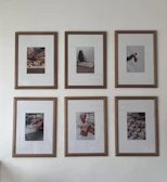 Six framed photographs hang on a wall in two rows of three. Each photograph captures different hands engaged in culinary activities, ranging from preparing dough to handling small pastries or desserts. The frames have a clean wooden border that complements the minimalist and artistic presentation of the photos.