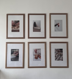 Six framed photographs hang on a wall in two rows of three. Each photograph captures different hands engaged in culinary activities, ranging from preparing dough to handling small pastries or desserts. The frames have a clean wooden border that complements the minimalist and artistic presentation of the photos.