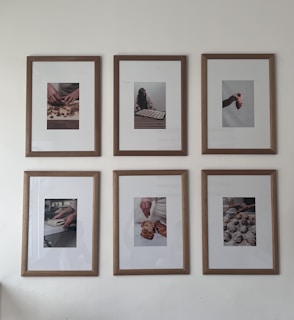 Six framed photographs hang on a wall in two rows of three. Each photograph captures different hands engaged in culinary activities, ranging from preparing dough to handling small pastries or desserts. The frames have a clean wooden border that complements the minimalist and artistic presentation of the photos.