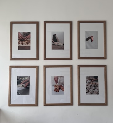 Six framed photographs hang on a wall in two rows of three. Each photograph captures different hands engaged in culinary activities, ranging from preparing dough to handling small pastries or desserts. The frames have a clean wooden border that complements the minimalist and artistic presentation of the photos.