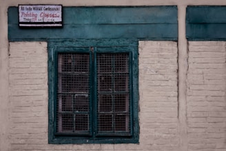 A rustic, weathered wall made of light-colored bricks contains a central window with a green frame. The window features mesh grids. Above, a small sign indicates information about painting classes organized by a women's conference.